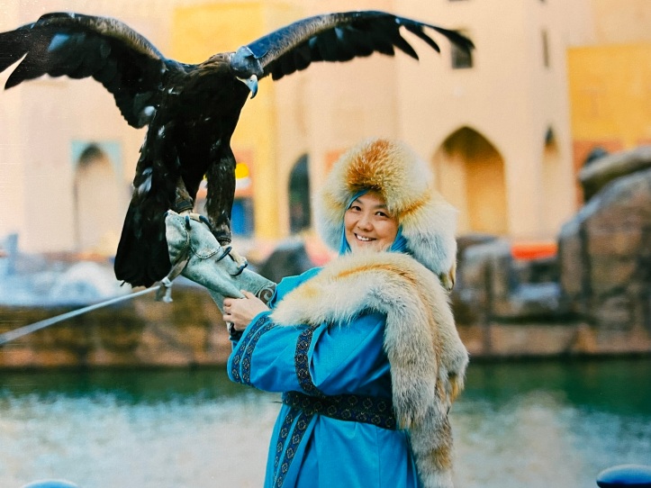 Author Fawzia Mai Tung, wearing a blue robe with a fur hood, smiling at the camera and posing with an eagle perched on her arm.